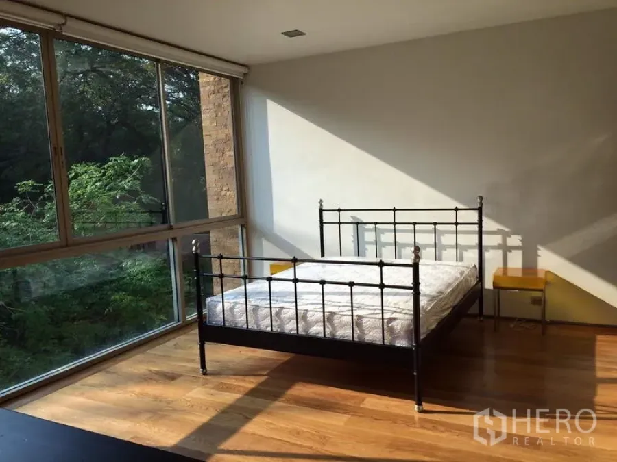 Condominium for rent in Khlong Toei, Bangkok - Sunlit bedroom with metal bedframe, hardwood floors, and full-height corner windows facing greenery.