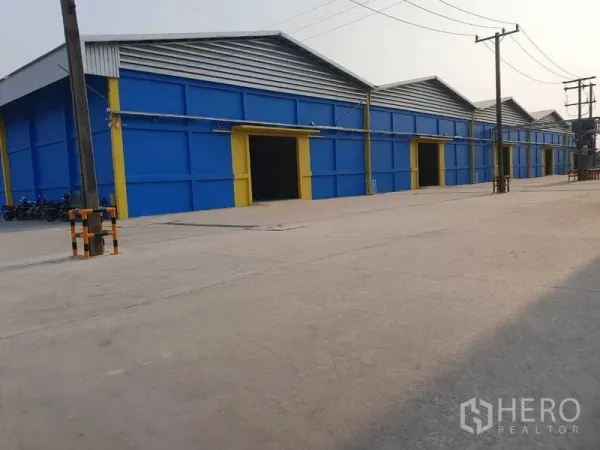 货栈 出租在Khlong Luang, Pathum Thani - 蓝黄相间的多联仓库外观，宽阔混凝土车道与电线杆。