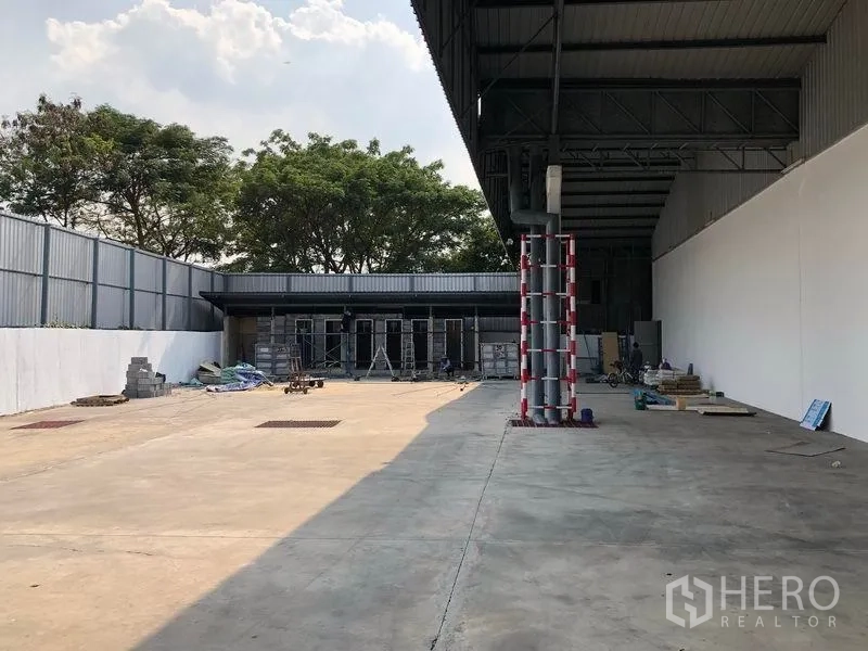 货栈 出租在Pak Kret, Nonthaburi - 宽敞露天院场，部分区域设雨棚，围栏完善，后方有树木。