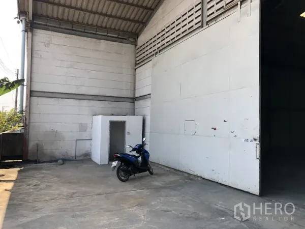 Warehouse for rent in Khlong Luang, Pathum Thani - Side loading area showing a large metal door, concrete yard and a small utility room beside the warehouse wall.