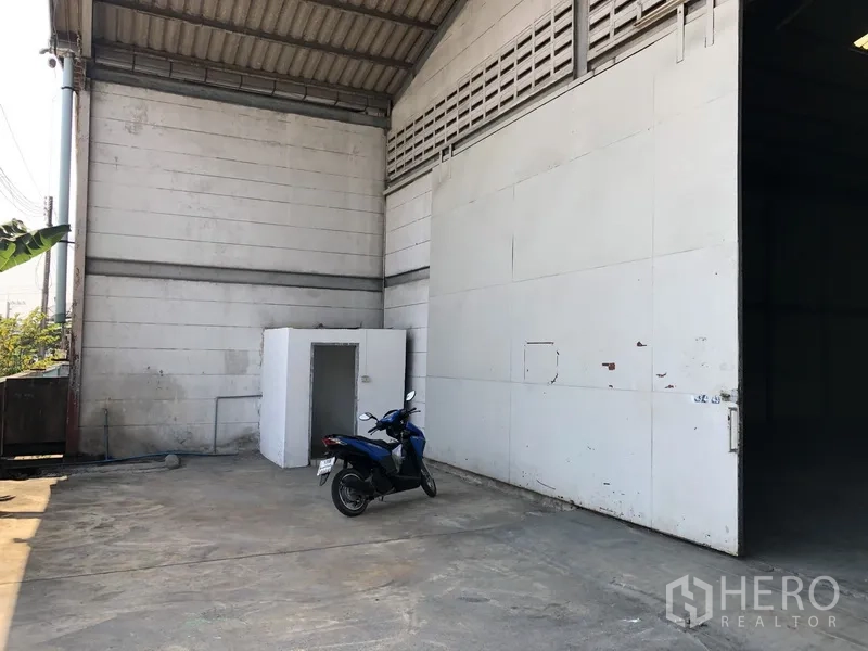 Warehouse for rent in Khlong Luang, Pathum Thani - Side loading area showing a large metal door, concrete yard and a small utility room beside the warehouse wall.