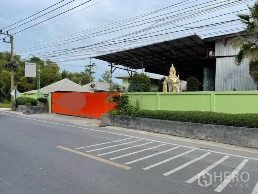 Warehouse for sale in Lam Luk Ka, Pathum Thani - Street-facing entrance of a secured warehouse with green boundary wall, bright orange gate, and steel-roofed buildings inside.
