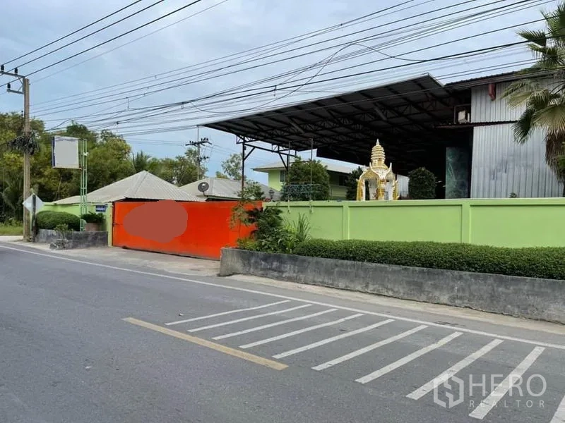 Warehouse for sale in Lam Luk Ka, Pathum Thani - Street-facing entrance of a secured warehouse with green boundary wall, bright orange gate, and steel-roofed buildings inside.