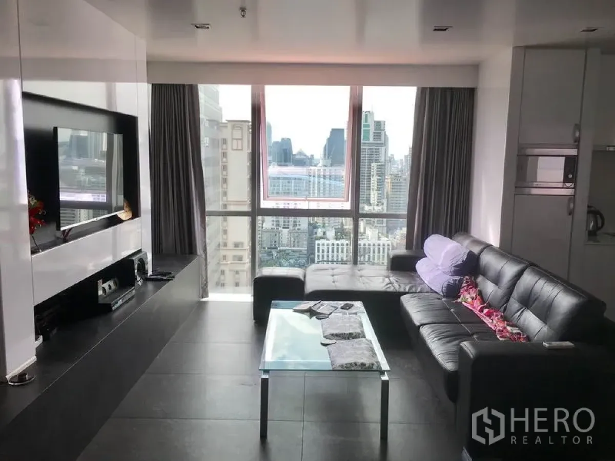 Condominium for rent in Watthana, Bangkok - Modern living room with black leather sofa, glass coffee table, and floor-to-ceiling windows overlooking the Bangkok skyline.