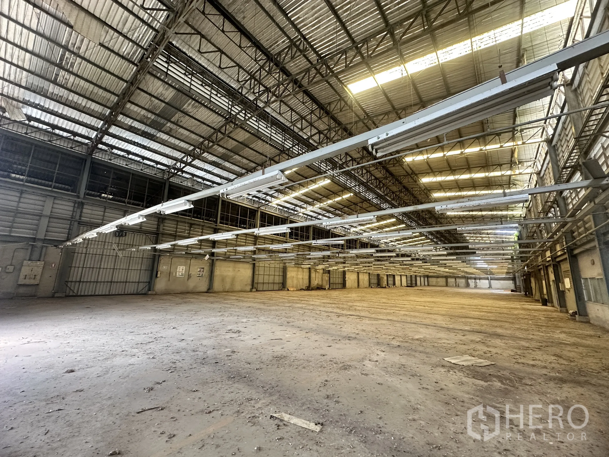 Factory for sale in Sai Noi, Nonthaburi - Long industrial bay with metal roofing, suspended lighting tracks, and wide gates.