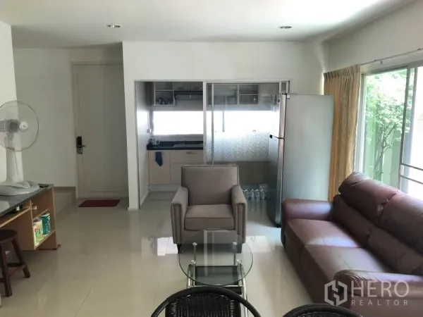 Townhouse for rent or sale in Suan Luang, Bangkok - Open-plan lounge with sofa set and glass coffee table facing a glass-partitioned kitchen and refrigerator.