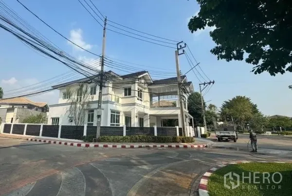 บ้าน ขายในPak Kret, Nonthaburi - มุมมองจากถนนของบ้านสีขาวสองชั้น พร้อมรั้วรอบแปลงมุม