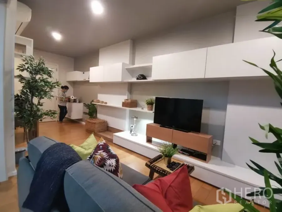 Condominium for rent or sale in Watthana, Bangkok - Modern living room with wall-mounted white storage, TV console and warm wood flooring.