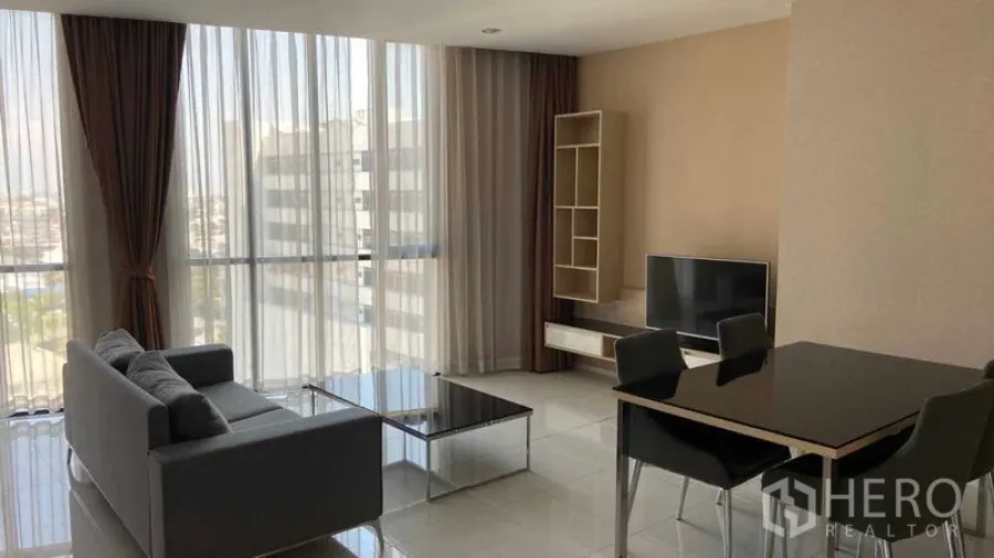 Condominium for rent in Watthana, Bangkok - Daylit living room with tall windows, sofa, TV stand, open shelving, and a four‑seat dining table.