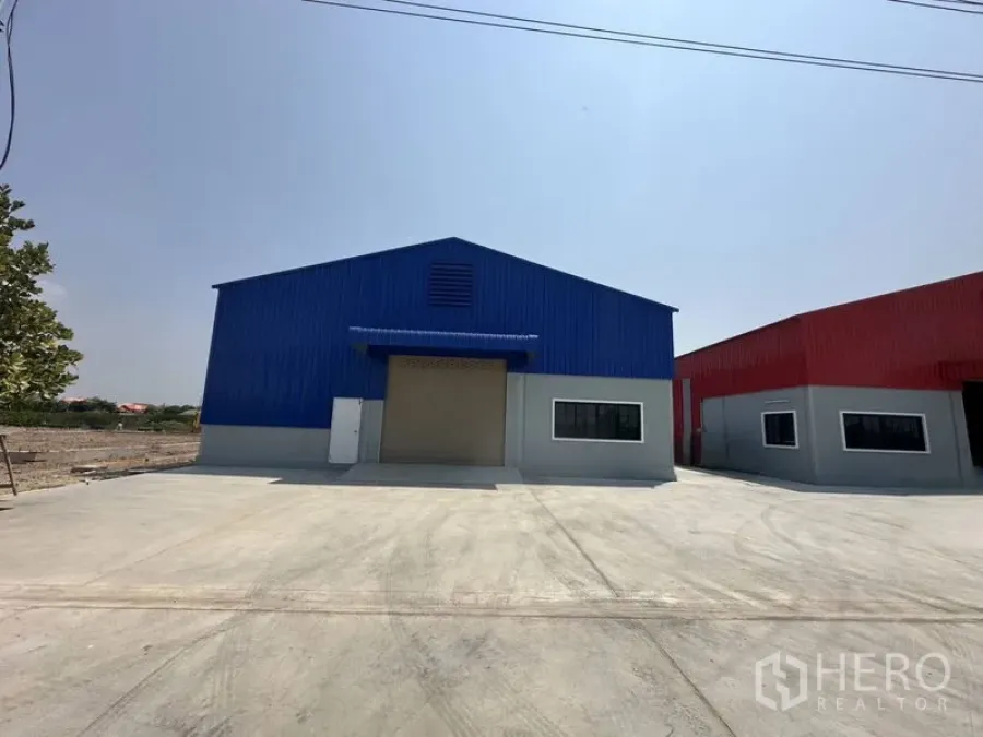 Warehouse for rent in Pak Kret, Nonthaburi - Front view of a new blue steel warehouse with a large roller shutter and wide concrete yard.