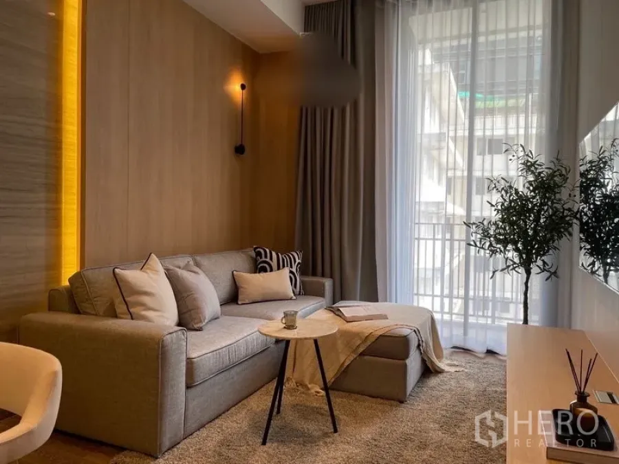 Condominium for rent or sale in Pathum Wan, Bangkok - Bright living room with grey sofa, ottoman, round coffee table and floor-to-ceiling sheer curtains opening to a balcony.
