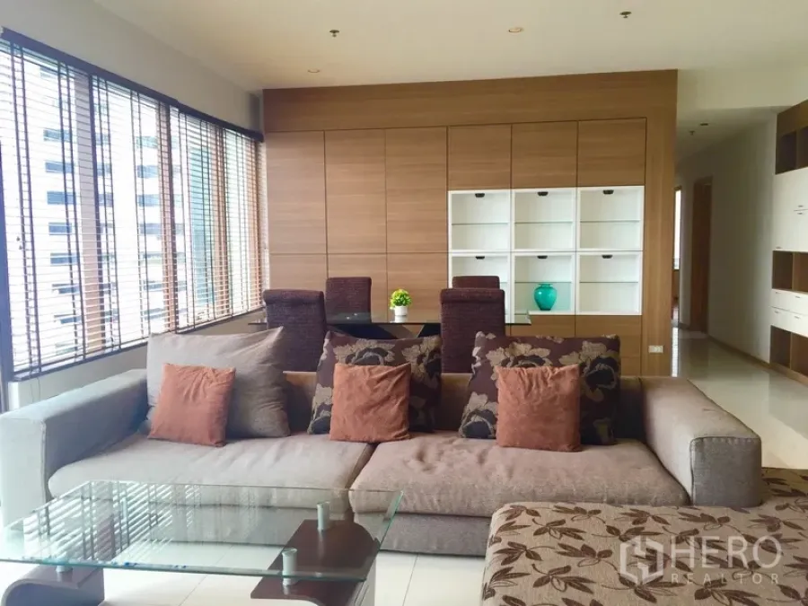 Condominium for rent in Khlong Toei, Bangkok - Bright living room with large sectional sofa, glass coffee table, dining area and wall of wooden cabinetry beside wide city-view windows.