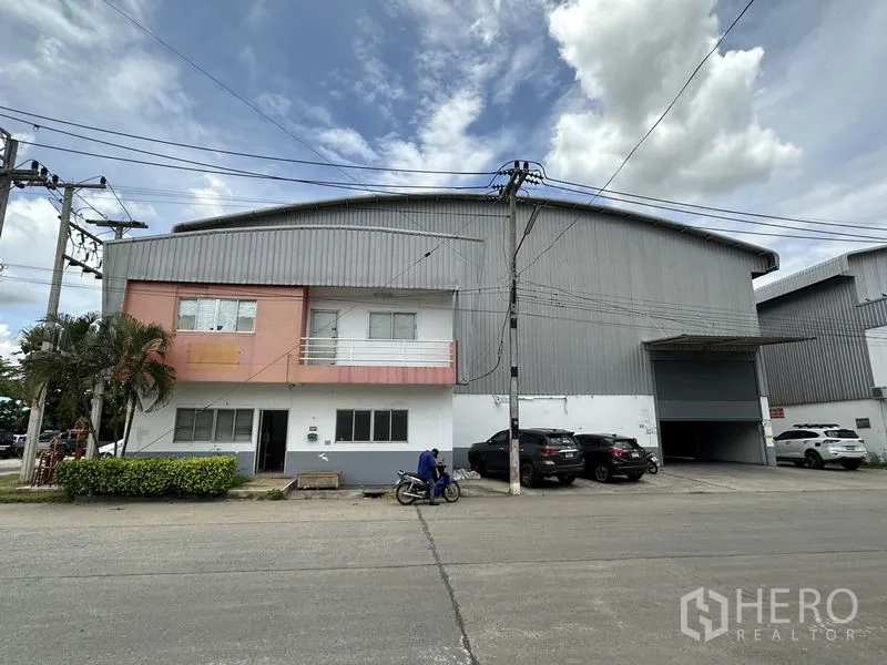 Factory for rent in Phra Nakhon Si Ayutthaya - Facade with office balcony, driveway and multiple parking spaces before the roller shutter entrance.