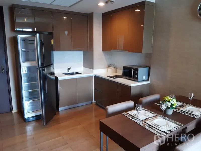 Condominium for rent in Khlong Toei, Bangkok - Modern L-shaped kitchen with glossy cabinets, full-size fridge, cooktop and a dining table in a 1-bedroom condo.