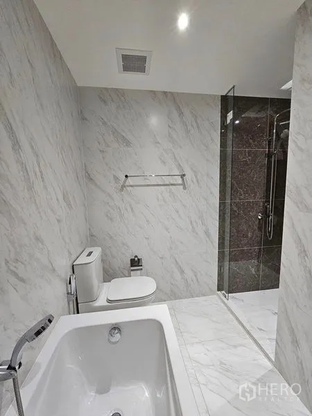 Condominium for rent or sale - Modern bathroom with white marble walls, bathtub, toilet, and glass shower.