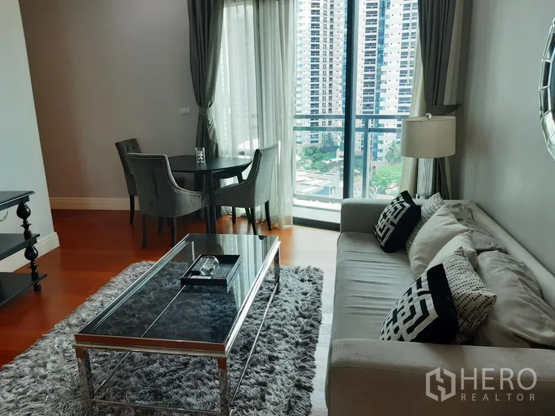 Condominium for rent or sale in Khlong Toei, Bangkok - Bright living room with grey sofa, glass coffee table, dining set and floor‑to‑ceiling windows to a balcony city view.