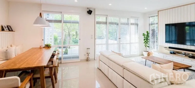 Detached House for rent - Bright open-plan dining and living area with long wooden table, white sofa, and wide windows with blinds.