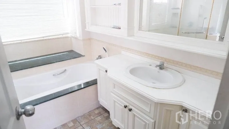 Detached House for rent in Prawet, Bangkok - White bathroom with vanity, mirror and a bathtub positioned under a window.