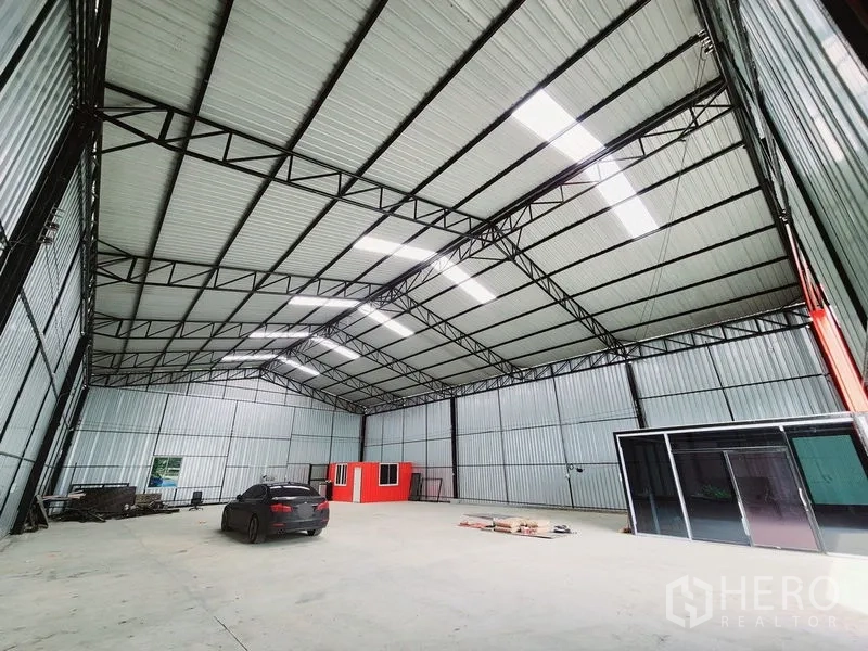 Warehouse for rent in Sam Phran, Nakhon Pathom - Spacious clear-span warehouse interior with steel trusses, skylights, concrete floor and a small red office box.