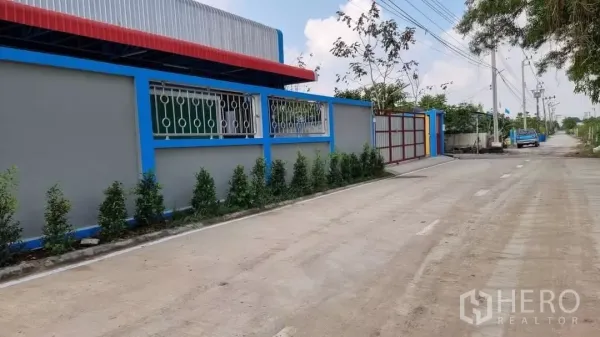 货栈 出租在Min Buri, Bangkok - 仓库侧立面与长围墙，金属窗格、绿化带及安静的街道与电线杆。
