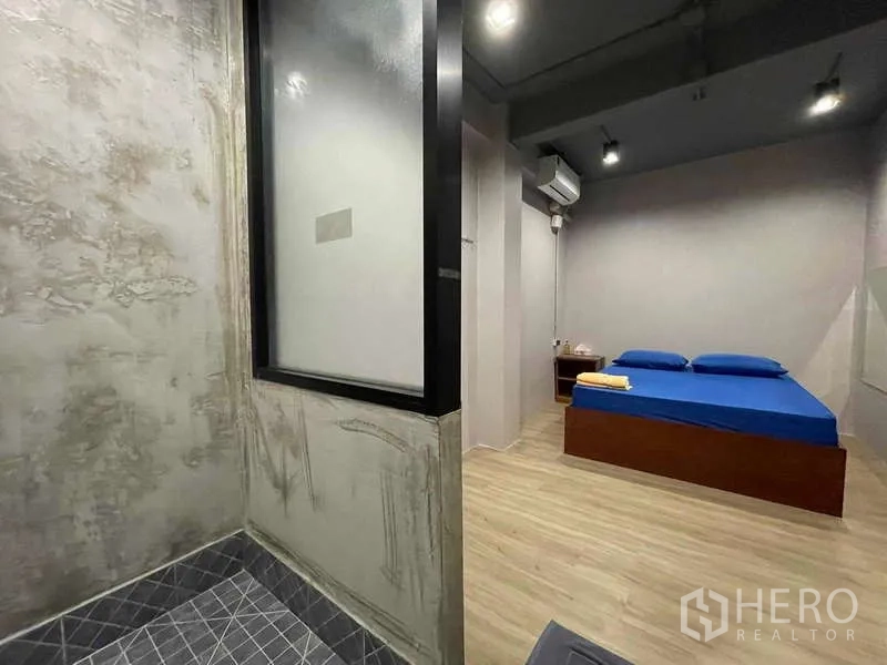 Shophouse for rent in Lat Phrao, Bangkok - Minimal bedroom with blue bedding, wood floor and adjacent concrete-look shower.