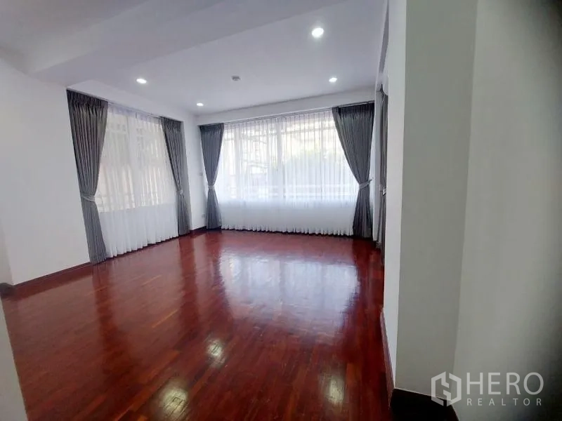 Condominium for rent in Watthana, Bangkok - Large empty room with wraparound windows, gray curtains, recessed lighting and wood flooring.