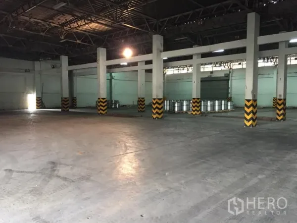 Warehouse for rent in Pathum Thani - Spacious warehouse interior with high steel-truss ceiling, concrete floor, and columns marked with yellow-black safety stripes.