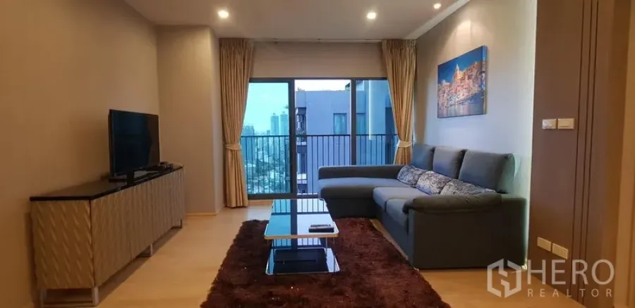 Condominium for rent in Khlong Toei, Bangkok - Bright living room with L-shaped sofa, TV console and balcony city view.