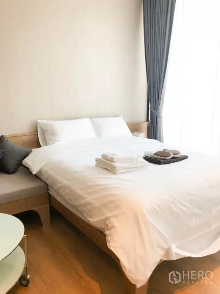 Condominium for rent in Khlong Toei, Bangkok - Bright bedroom with double bed, grey curtains and a side bench by the window.