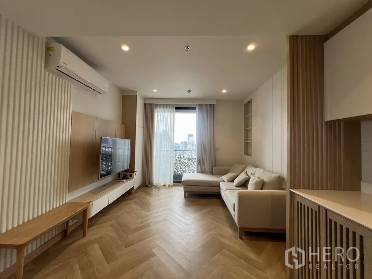 Condominium for rent or sale in Chatuchak, Bangkok - Minimalist living room with L‑shaped sofa, wall‑mounted TV, balcony door and city view over herringbone wood floors.