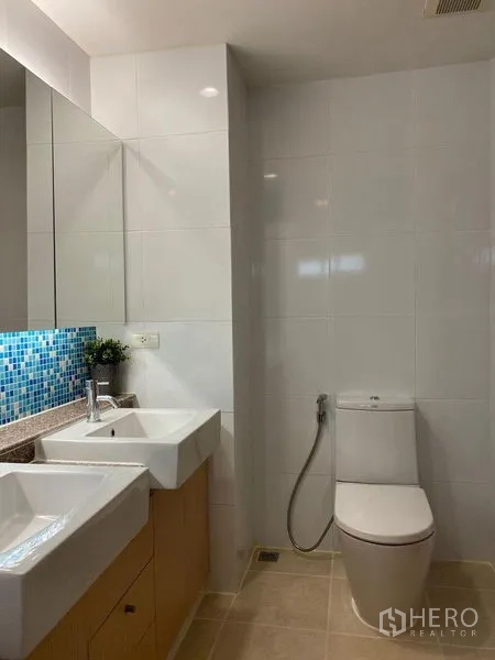 Condominium for sale in Phra Khanong, Bangkok - Modern bathroom with double sinks, large mirror, toilet, and white wall tiles.