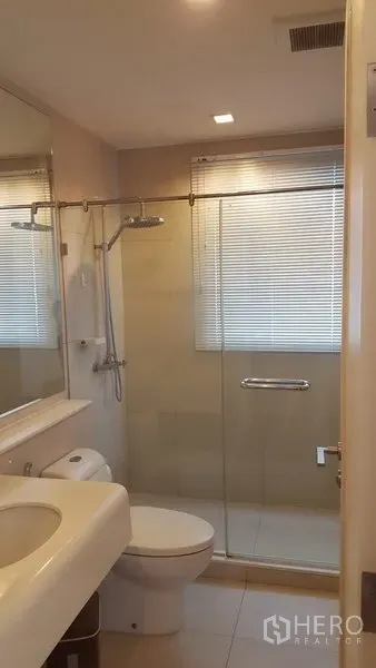Condominium for rent in Bang Rak, Bangkok - Modern bathroom with glass-enclosed shower, vanity and toilet under window blinds.