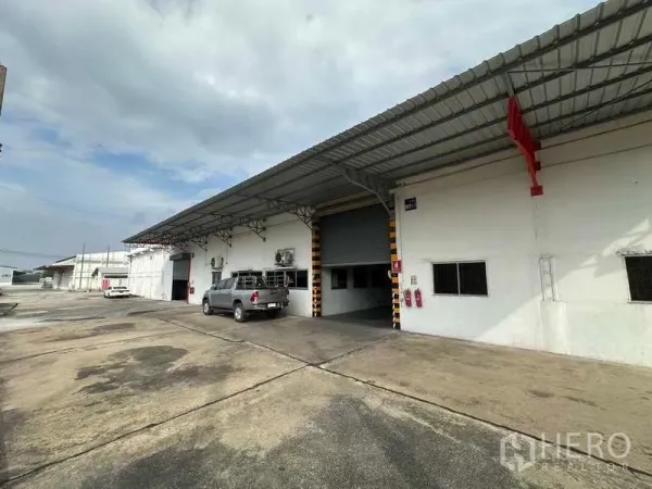 货栈 出租在Mueang Pathum Thani, Pathum Thani - 邦卡迪园区内带雨棚的装卸区与卷闸门仓库外观。