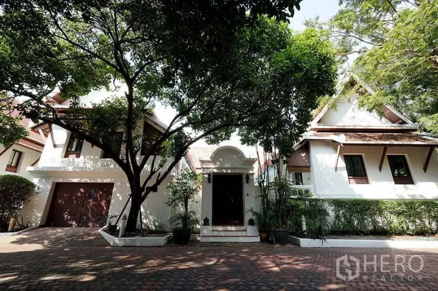 Detached House for rent in Watthana, Bangkok - Front exterior of a Thai-inspired detached house with garage, mature trees, and a brick-paved driveway.