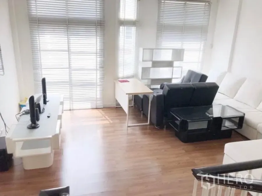 Townhouse for rent in Prawet, Bangkok - Bright living area with large windows, blinds, wood flooring, black and white sofas, and a work desk.