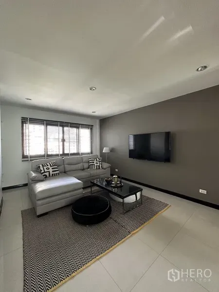 Condominium for rent in Khlong Toei, Bangkok - Contemporary living room with gray sectional sofa, wall-mounted TV, and window.
