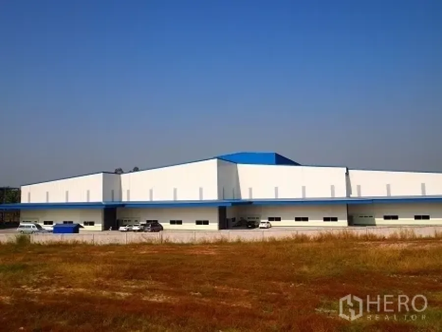 Factory for rent in Si Racha, Chon Buri - Wide exterior view of a low-rise white factory with blue roof accents and parking, seen across a field.