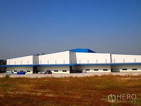 Factory for rent in Si Racha, Chon Buri - Wide exterior view of a low-rise white factory with blue roof accents and parking, seen across a field.