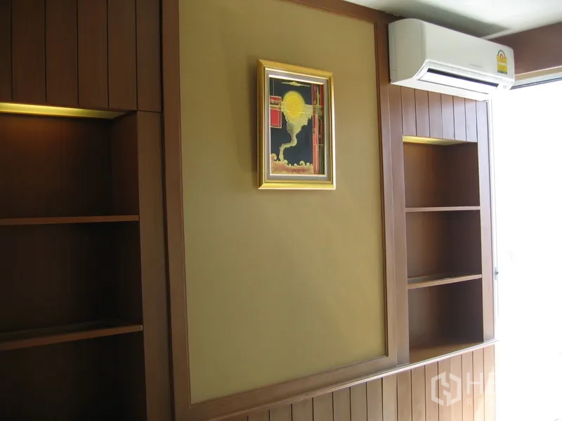 Condominium for rent in Khlong Toei, Bangkok - Bedroom wall with built-in shelves, air conditioner and framed artwork near a bright window.