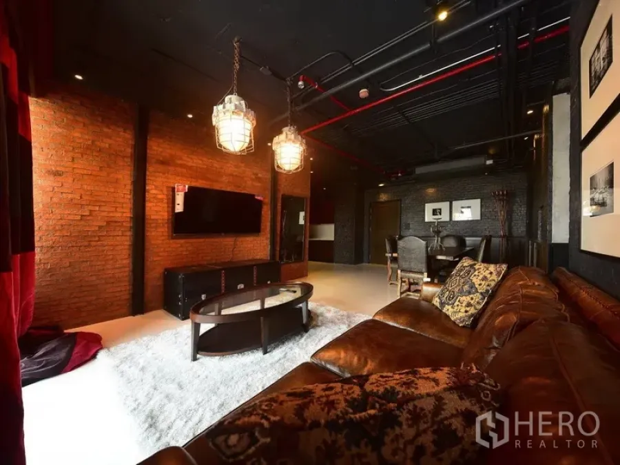 Condominium for rent or sale in Khlong Toei, Bangkok - Industrial loft-style living room with exposed brick walls, leather sofa, pendant lights and a wall-mounted TV in a Sukhumvit condo.