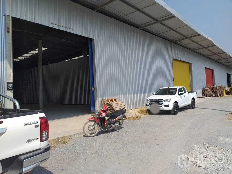 Warehouse for rent in Pak Kret, Nonthaburi - Covered loading area with multiple colored roller doors and parking for pickup trucks.