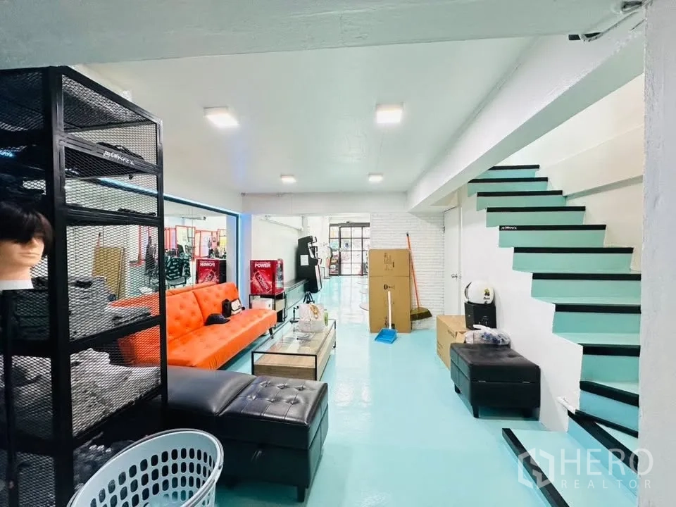 Shophouse for sale in Watthana, Bangkok - Open ground floor showing orange sofa, glass entrance ahead, and teal staircase to upper level.