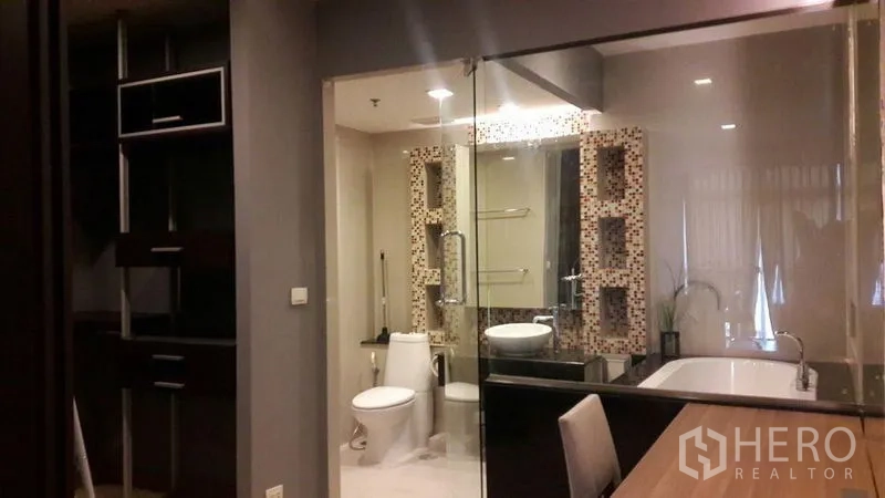 Condominium for rent in Khlong Luang, Pathum Thani - Bedroom view into a glass-partitioned bathroom showing tub, toilet and vanity with mosaic shelving.