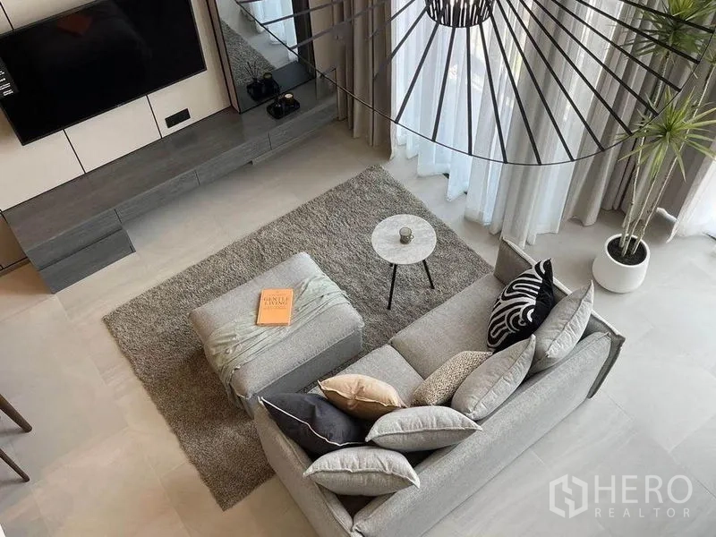 Condominium for rent in Pathum Wan, Bangkok - Top view of airy living area with grey sofa set, ottoman on rug, round side table and full-height windows.