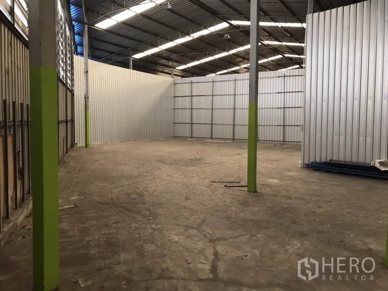 Warehouse for rent in Bang Na, Bangkok - Open warehouse space with tall ceiling and green-marked support posts beside corrugated partitions.
