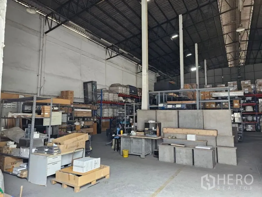 Warehouse for rent in Khlong Toei, Bangkok - High-ceiling warehouse interior with steel truss roof, shelving racks, concrete floor and workbench area.