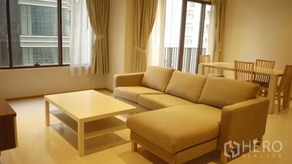 Condominium for rent in Khlong Toei, Bangkok - Open-plan lounge and dining area with sofa, white coffee table, wooden dining set and balcony doors facing the city.