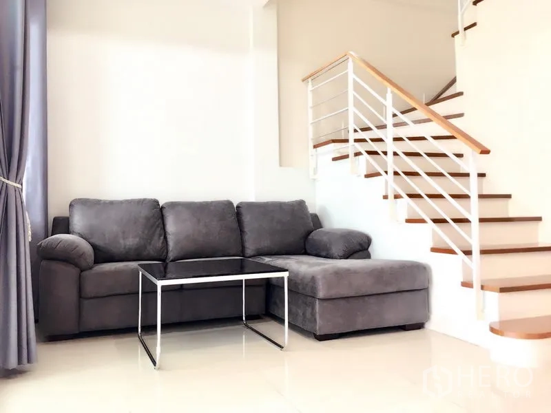 Townhouse for rent in Prawet, Bangkok - A bright living room with a gray L-shaped sofa beside a wooden staircase.