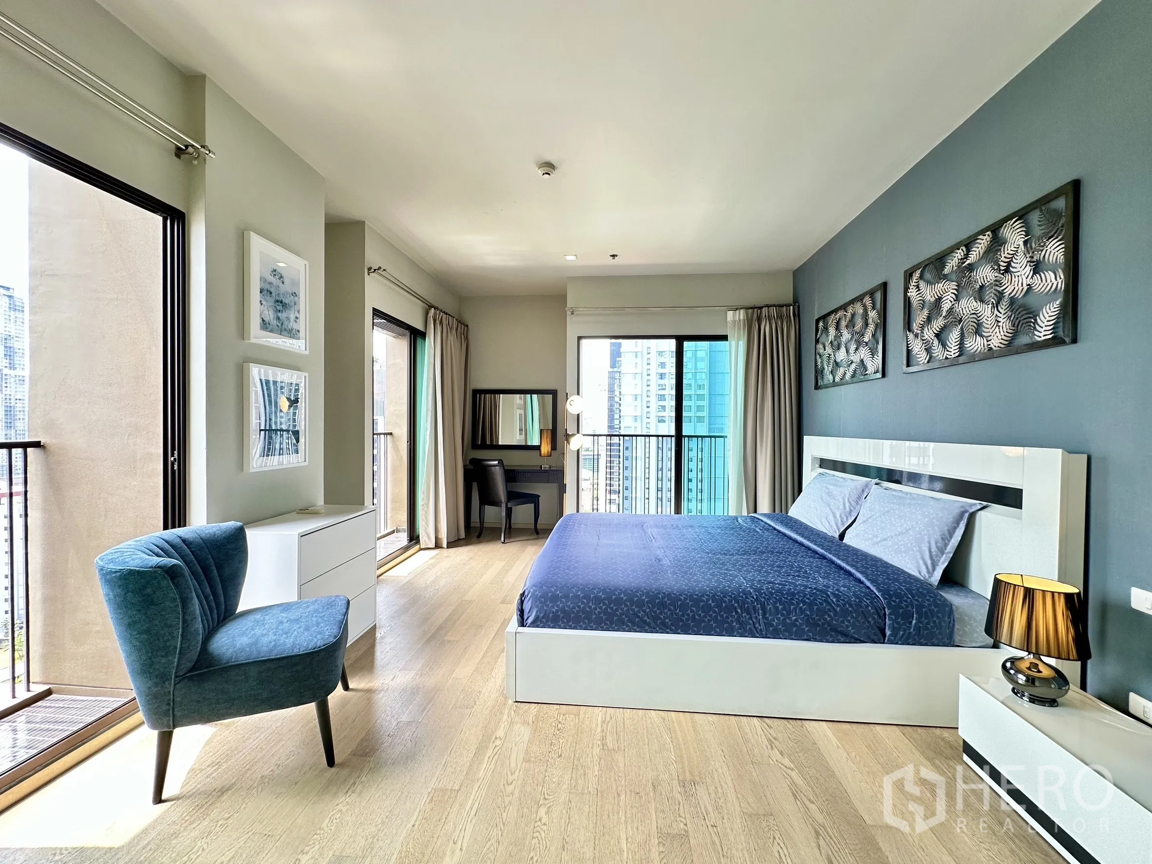 Condominium for rent in Bangkok - Corner bedroom with two balcony doors, blue armchair and abundant natural light.