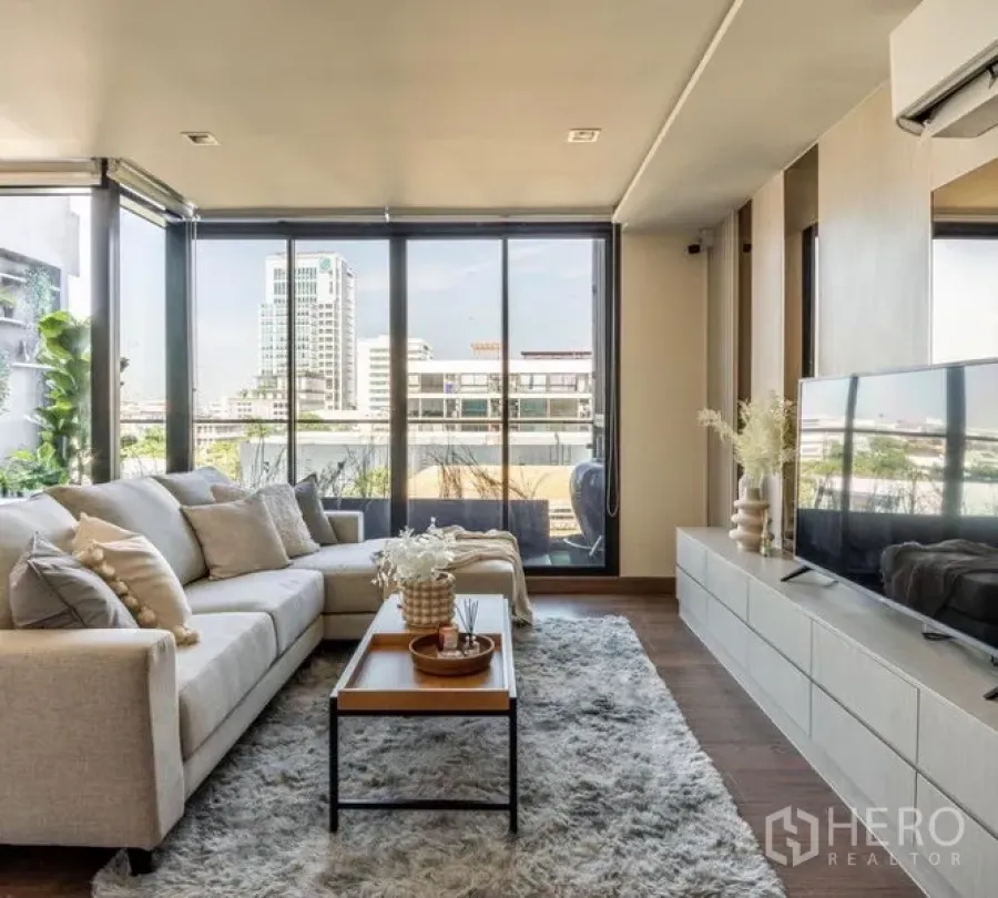 Condominium for rent or sale in Phra Khanong, Bangkok - Bright living room with beige sectional sofa, shag rug, TV console and floor-to-ceiling windows opening to a balcony with city views.